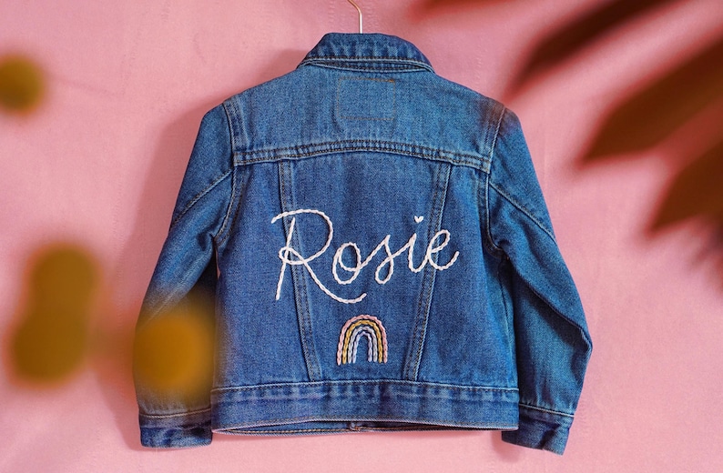 May include: A blue denim jacket with the name "Rosie" embroidered on the back in white thread. A rainbow is embroidered below the name.