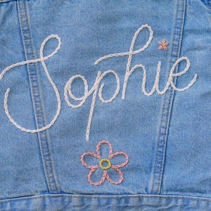 Puede incluir: Una chaqueta vaquera azul claro con el nombre "Sophie" bordado en hilo blanco. Una flor rosa y amarilla est&aacute; bordada debajo del nombre.