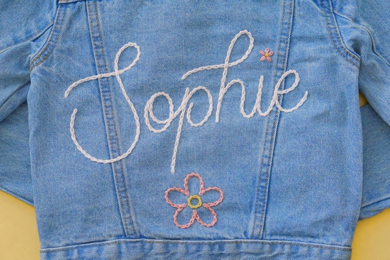 Puede incluir: Una chaqueta vaquera azul claro con el nombre "Sophie" bordado en hilo blanco. Una flor rosa y amarilla est&aacute; bordada debajo del nombre.