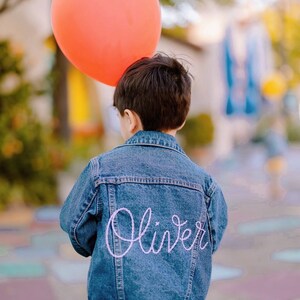 Puede incluir: Una persona joven lleva una chaqueta vaquera azul con el nombre "Oliver" bordado en la espalda con hilo rosa. La persona sostiene un globo naranja brillante.