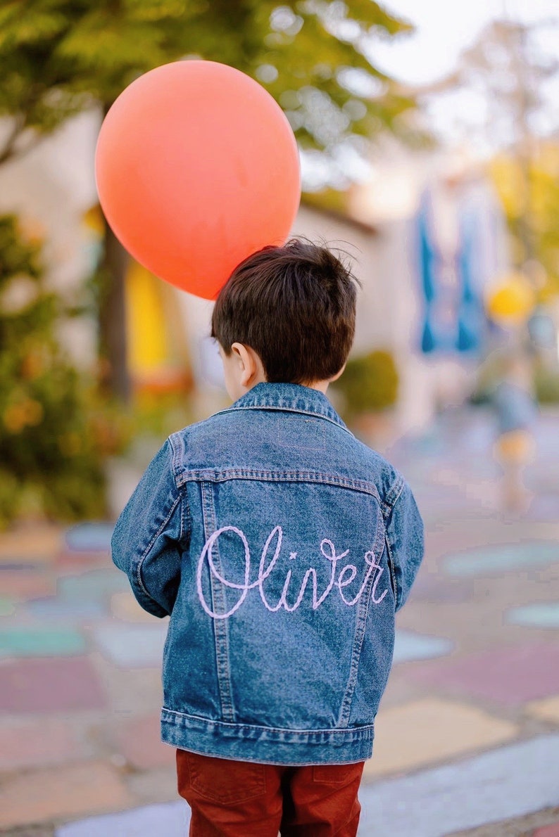 Puede incluir: Una persona joven lleva una chaqueta vaquera azul con el nombre "Oliver" bordado en la espalda con hilo rosa. La persona sostiene un globo naranja brillante.