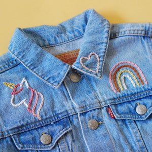 May include: A blue denim jacket with embroidered designs, including a unicorn, a rainbow, and a heart on the collar.
