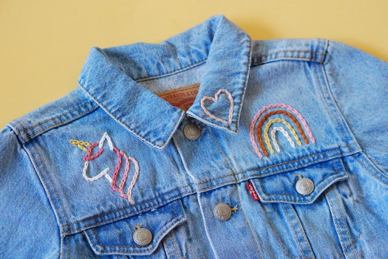 May include: A blue denim jacket with embroidered designs, including a unicorn, a rainbow, and a heart on the collar.