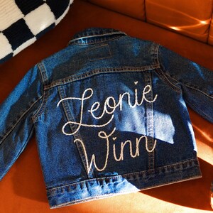 Puede incluir: Una chaqueta vaquera azul con bordado blanco que dice "Leonie Winn" en la espalda.