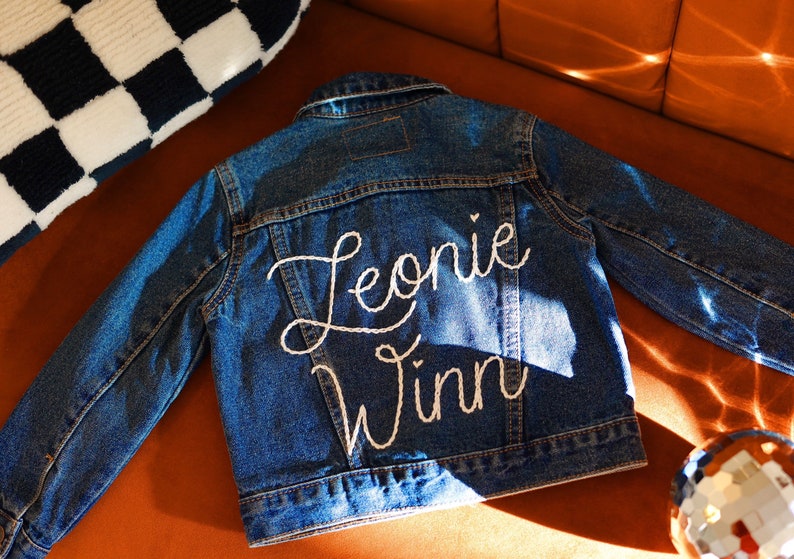 Puede incluir: Una chaqueta vaquera azul con bordado blanco que dice "Leonie Winn" en la espalda.