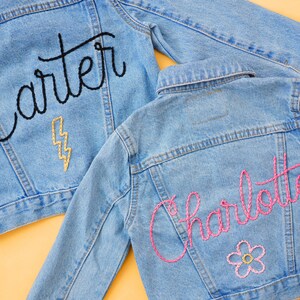 Puede incluir: Dos chaquetas vaqueras con nombres bordados. Una chaqueta tiene el nombre "Carter" bordado en hilo negro con un rayo dorado. La otra chaqueta tiene el nombre "Charlotte" bordado en hilo rosa con una flor rosa.