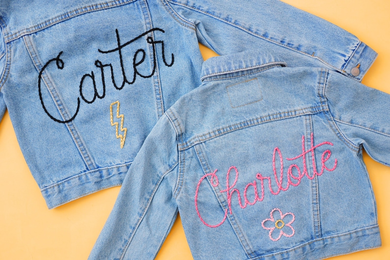 Puede incluir: Dos chaquetas vaqueras con nombres bordados. Una chaqueta tiene el nombre "Carter" bordado en hilo negro con un rayo dorado. La otra chaqueta tiene el nombre "Charlotte" bordado en hilo rosa con una flor rosa.