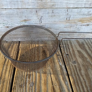 May include: A rusty metal wire mesh strainer with a single handle. The strainer is sitting on a wooden surface.
