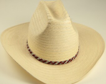 Könnte beinhalten: Hellbeige Cowboyhut aus Stroh mit breiter Krempe. Der Hut hat ein dekoratives Band mit einem Muster aus Rot, Blau und Weiß. Der Hut dient als Sonnenschutz. Der Hut hat einen Krempen-Durchmesser von ca. 30 cm.