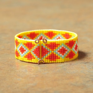 May include: A yellow, red, and white beaded bracelet with a geometric pattern. The bracelet has two copper clasps.