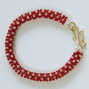 May include: A red and clear beaded bracelet with a gold clasp. The bracelet is made with small, round beads that are arranged in a spiral pattern.