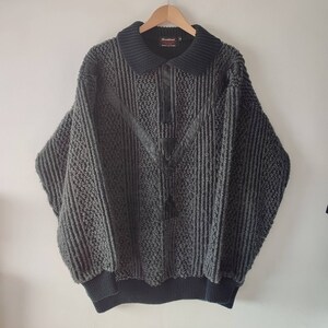 May include: A gray and black vintage sweater with a textured knit pattern. The sweater features a collared neckline, a V-shaped leather accent, and three buttons. The sleeves are long and the bottom hem is ribbed.