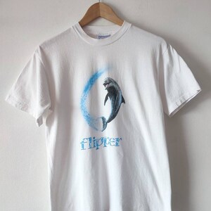 May include: White t-shirt featuring a blue and white graphic of a dolphin leaping from the water. The word "Flipper" is printed beneath the dolphin.