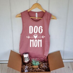 May include: A dusty rose tank top with the words "DOG MOM" in white, hanging on a wooden hanger. The tank top is inside an open cardboard box with a rose gold tumbler, a small plant, and decorative filler.