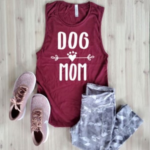 May include: A maroon tank top with the text "DOG MOM" in white, a pair of gray patterned leggings, and rose gold athletic shoes with white soles and laces. The items are arranged on a light wood surface.