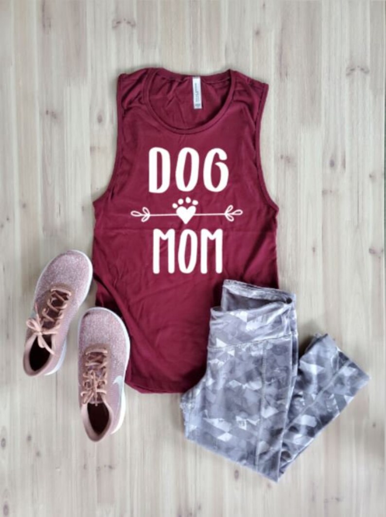 May include: A maroon tank top with the text "DOG MOM" in white, a pair of gray patterned leggings, and rose gold athletic shoes with white soles and laces. The items are arranged on a light wood surface.