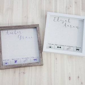 K&ouml;nnte beinhalten: Zwei Holz-Schattenboxen mit wei&szlig;en Passepartouts und Glasfronten. Die obere Box tr&auml;gt den Namen "Elijah Aaron" in Kursivschrift. Die untere Box tr&auml;gt den Namen "Amber Grace" in Kursivschrift. Beide Boxen enthalten Geburtsdaten wie Uhrzeit, Datum, L&auml;nge und Gewicht.