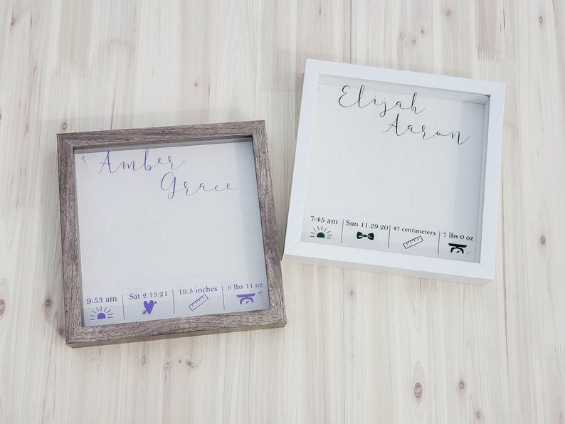 K&ouml;nnte beinhalten: Zwei Holz-Schattenboxen mit wei&szlig;en Passepartouts und Glasfronten. Die obere Box tr&auml;gt den Namen "Elijah Aaron" in Kursivschrift. Die untere Box tr&auml;gt den Namen "Amber Grace" in Kursivschrift. Beide Boxen enthalten Geburtsdaten wie Uhrzeit, Datum, L&auml;nge und Gewicht.