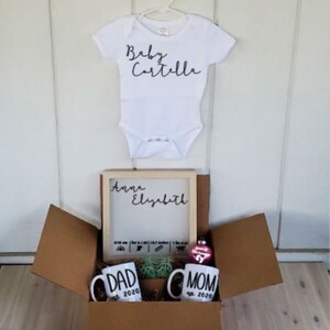 K&ouml;nnte beinhalten: Eine braune Box mit einem wei&szlig;en Strampler, auf dem "Baby Cartelle" steht. Die Box enth&auml;lt au&szlig;erdem ein gerahmtes Schild mit der Aufschrift "Anna Elizabeth", zwei Tassen mit der Aufschrift "Papa est. 2020" und "Mama est. 2020" sowie einen rosa Anh&auml;nger.