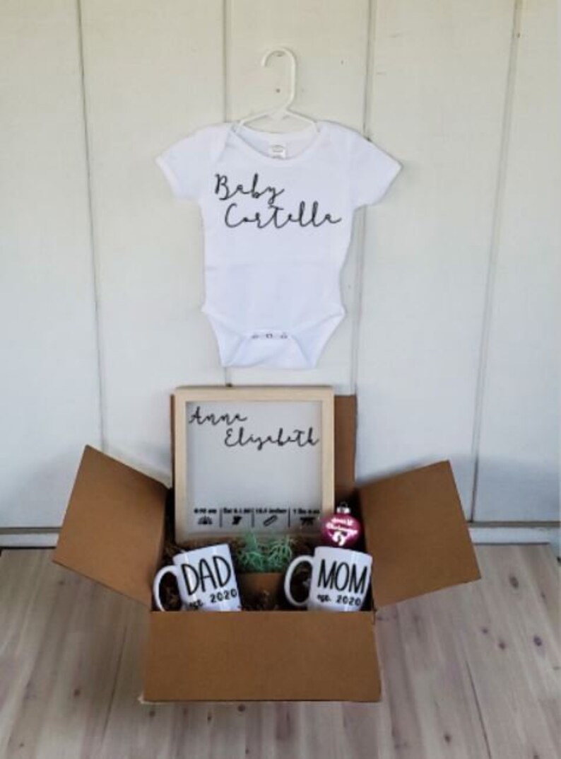 K&ouml;nnte beinhalten: Eine braune Box mit einem wei&szlig;en Strampler, auf dem "Baby Cartelle" steht. Die Box enth&auml;lt au&szlig;erdem ein gerahmtes Schild mit der Aufschrift "Anna Elizabeth", zwei Tassen mit der Aufschrift "Papa est. 2020" und "Mama est. 2020" sowie einen rosa Anh&auml;nger.