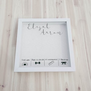 K&ouml;nnte beinhalten: Ein wei&szlig;er Bilderrahmen mit einem wei&szlig;en Passepartout und einem wei&szlig;en Hintergrund. Der Rahmen enth&auml;lt den Namen "Elijah Aaron" in schwarzer Kursivschrift. Unter dem Namen befindet sich eine Textzeile mit der Aufschrift "7:45 Uhr | So 11.29.20 | 47 Zentimeter | 7 lbs 0 oz". Unter dem Text befinden sich vier Symbole: eine Sonne, eine Fliege, ein Lineal und eine Waage.