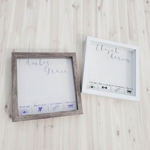 K&ouml;nnte beinhalten: Zwei wei&szlig;e Schattenboxen mit braunen Holzrahmen. Die obere Box tr&auml;gt die Namen "Elijah Aaron" in lilafarbener Kursivschrift. Die untere Box tr&auml;gt die Namen "Amber Grace" in lilafarbener Kursivschrift. Beide Boxen haben ein lilafarbenes Sonnen-, Herz-, Lineal- und Waagen-Symbol mit den entsprechenden Geburtsdaten.