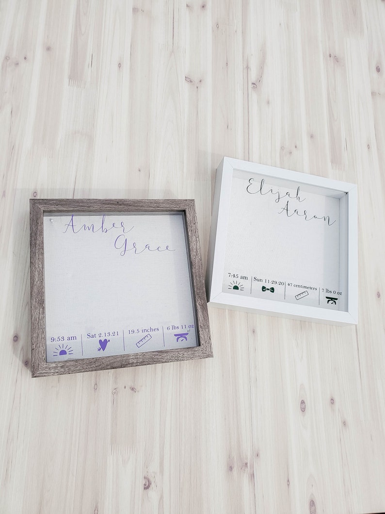 K&ouml;nnte beinhalten: Zwei wei&szlig;e Schattenboxen mit braunen Holzrahmen. Die obere Box tr&auml;gt die Namen "Elijah Aaron" in lilafarbener Kursivschrift. Die untere Box tr&auml;gt die Namen "Amber Grace" in lilafarbener Kursivschrift. Beide Boxen haben ein lilafarbenes Sonnen-, Herz-, Lineal- und Waagen-Symbol mit den entsprechenden Geburtsdaten.