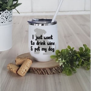 May include: White insulated wine tumbler with a black lid and a white straw. The tumbler has the text "I just want to drink wine & pet my dog" with a gray dog silhouette. Corks and greenery are in the foreground.