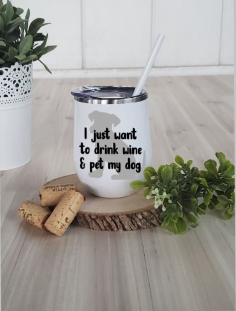 May include: White insulated wine tumbler with a black lid and a white straw. The tumbler has the text "I just want to drink wine & pet my dog" with a gray dog silhouette. Corks and greenery are in the foreground.