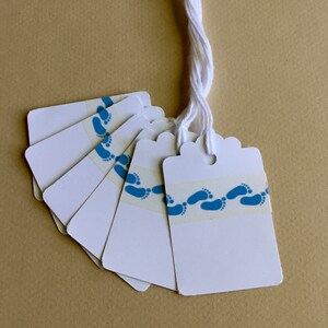 May include: A set of ten white gift tags with blue footprints printed on them. The tags are attached to a white string.