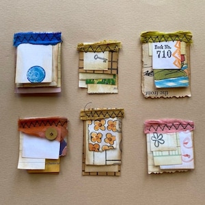 May include: Five small handmade paper journals with different fabric covers and decorative elements. The covers include a blue fabric with a stitched border, a yellow fabric with a stitched border, a brown fabric with a stitched border, a yellow fabric with a stitched border, and a pink fabric with a stitched border. The journals are decorated with various paper scraps, including a key, a flower, a circle, and a book number.