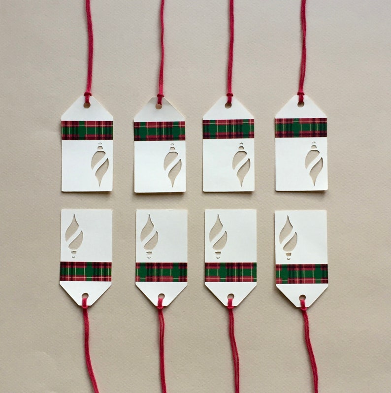 May include: Eight white gift tags with red and green plaid ribbon and a cut-out ornament design. The tags are tied with red string.