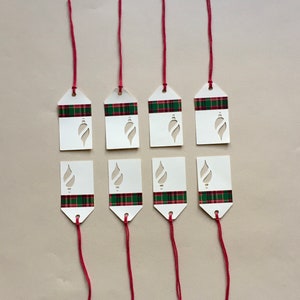 May include: Eight white gift tags with a red and green plaid pattern and a cut-out ornament design. The tags are attached to red strings.