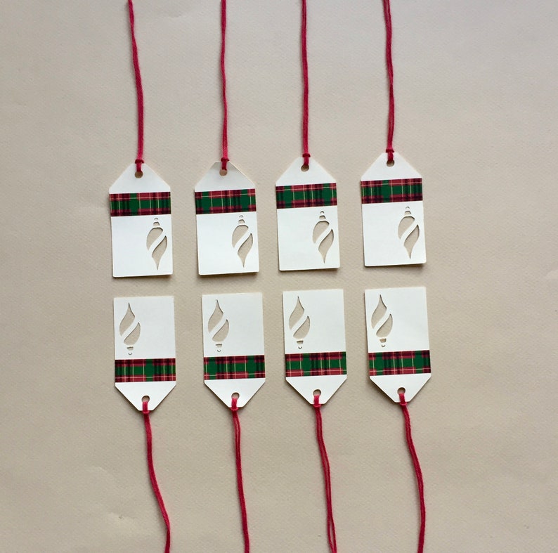 May include: Eight white gift tags with a red and green plaid pattern and a cut-out ornament design. The tags are attached to red strings.