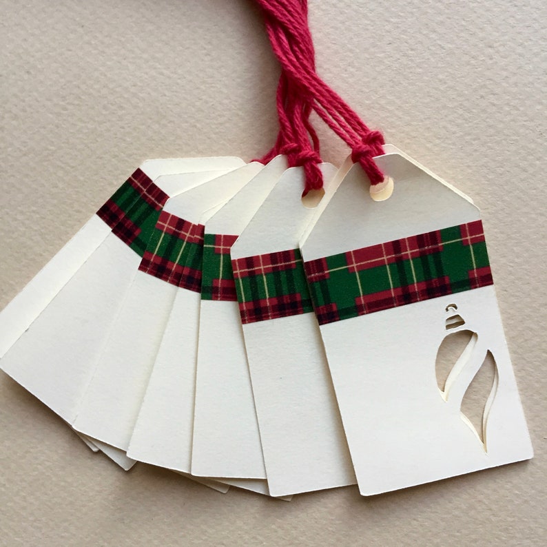 May include: A set of eight white gift tags with red and green plaid ribbon and a cut-out ornament design. The tags are tied together with red string.