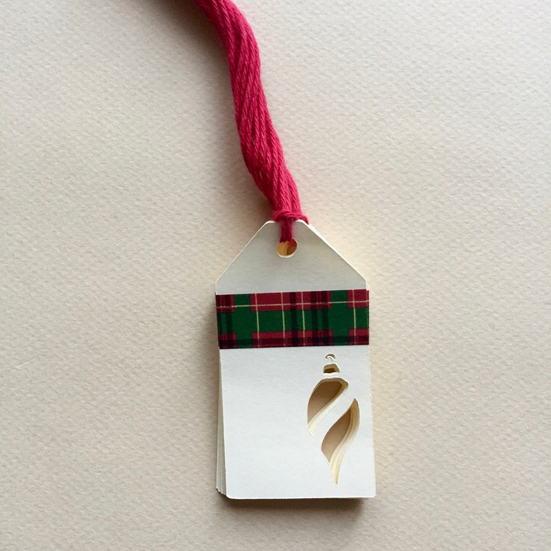 May include: White gift tag with a red and green plaid ribbon and a cut-out ornament shape. The tag is attached to a red string.