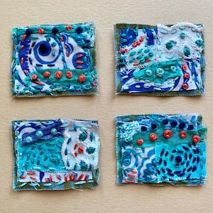 May include: Four small, square textile art pieces. Each features a unique design with blue, teal, and white fabric, accented with red and teal embroidery. The edges are finished with a green border, creating a cohesive set.