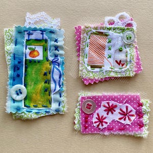 May include: Three handmade fabric ornaments with a variety of textures and colors. The first ornament is blue and green with a white button and a small orange fruit. The second ornament is pink and green with a white button and a red heart. The third ornament is pink with white polka dots and red embroidered flowers.