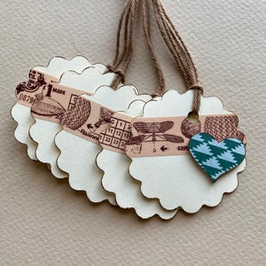May include: Five white scalloped paper gift tags with twine attached. Each tag has a different vintage design, including a dragonfly, a heart, and a grid pattern. The tags are perfect for adding a touch of rustic charm to gifts.