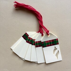May include: A set of eight white gift tags with a green and red plaid ribbon and a cut-out ornament design. The tags are tied together with a red string.