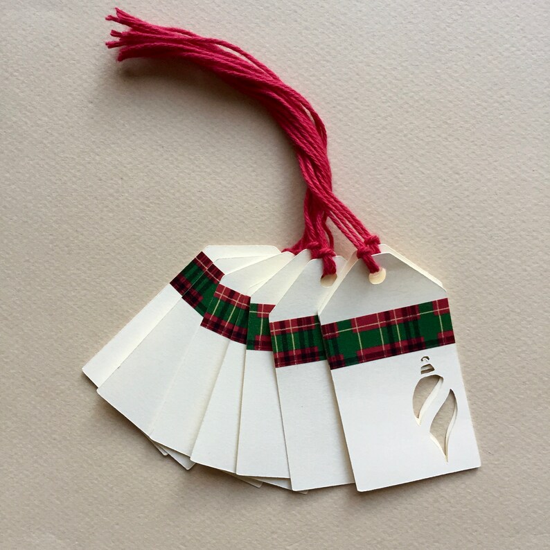 May include: A set of eight white gift tags with a green and red plaid ribbon and a cut-out ornament design. The tags are tied together with a red string.