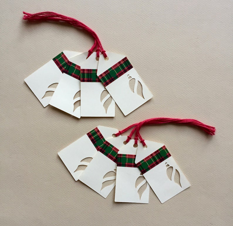 May include: Eight white gift tags with red and green plaid ribbon and a cut-out design of a leaf. The tags are tied with red string.