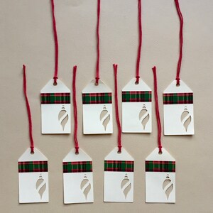 May include: Eight white gift tags with a red and green plaid pattern and a cut-out ornament shape. The tags are tied with red string.