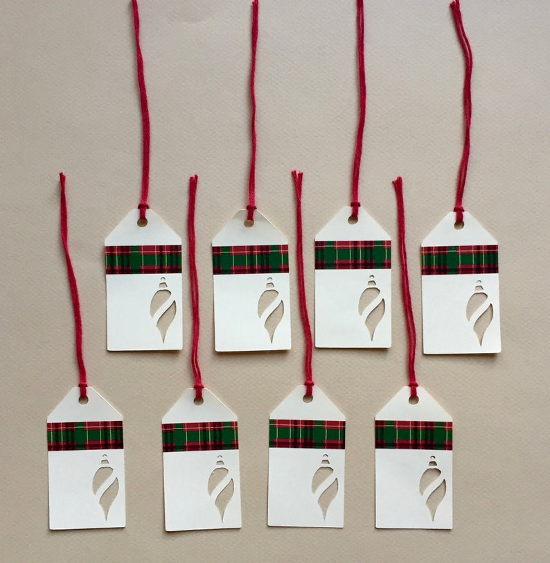 May include: Eight white gift tags with a red and green plaid pattern and a cut-out ornament shape. The tags are tied with red string.