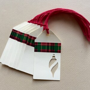 May include: A set of ten white gift tags with a red and green plaid ribbon and a cut-out ornament shape. The tags have a hole punched at the top and come with red string.