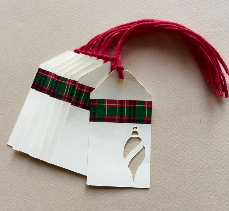 May include: A set of ten white gift tags with a red and green plaid ribbon and a cut-out ornament shape. The tags have a hole punched at the top and come with red string.