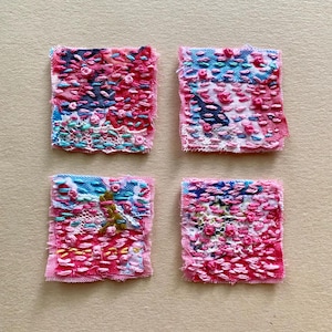 May include: Four small square fabric patches with pink, blue, and white embroidery. The patches are stitched together with a variety of stitches and patterns.