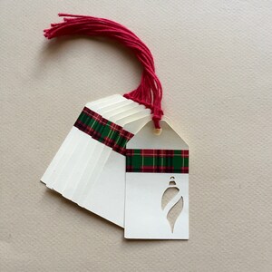 May include: A stack of white gift tags with a red and green plaid ribbon and a red tassel. The tags have a cut-out design of a Christmas ornament.