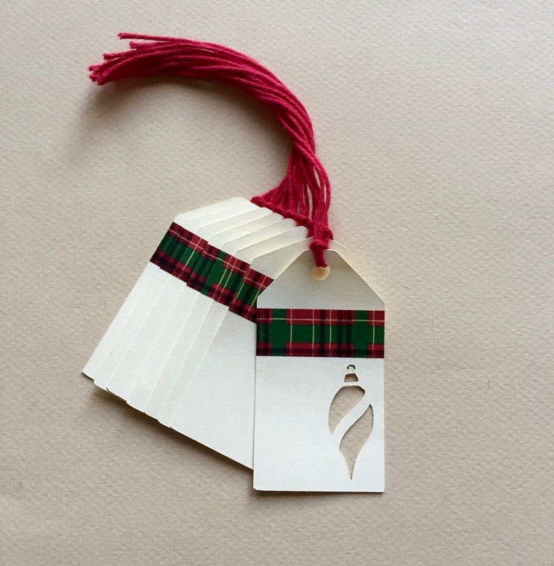 May include: A stack of white gift tags with a red and green plaid ribbon and a red tassel. The tags have a cut-out design of a Christmas ornament.