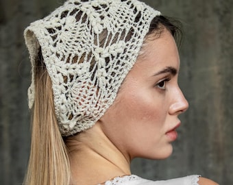 May include: Cream-colored, open-weave crochet head covering. The head covering is worn on the head and tied at the back. The head covering is made of a soft, delicate yarn. The background is a muted gray.